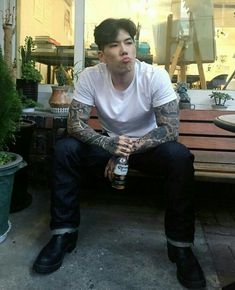 a man with tattoos sitting on a wooden bench holding a beer in one hand and looking at the camera