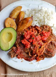 a white plate topped with meat, rice and veggies next to an avocado