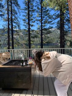 a woman bending over to look at an outdoor fire pit on a deck with pine trees in the background