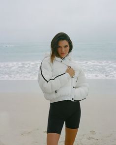 a woman standing on top of a sandy beach next to the ocean wearing a white jacket and black shorts