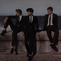 three young men in suits sitting on a wall near the ocean