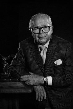 an old man in a suit and tie leaning on a table with his arms crossed