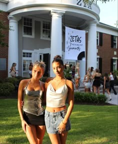 two young women standing in front of a building with people walking around the lawn behind them