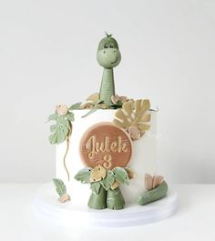 a white cake with a green dinosaur on top and leaves around the edges that says gulo's