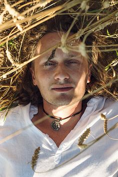 a man with long hair wearing a white shirt and looking at the camera through some grass