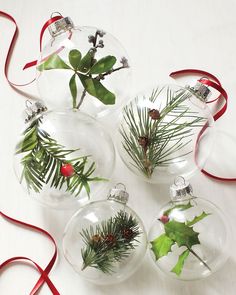 four glass ornaments with holly and red ribbon hanging from them on a white table top