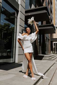 a woman in a white dress is posing on the street with her arms up and flowers in her hair