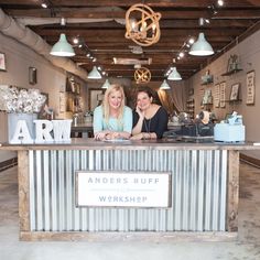 two women sitting at a counter in an artisan shop, smiling for the camera