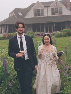 a man and woman standing in front of a large house holding wine glasses while walking through tall grass