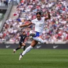 a woman kicking a soccer ball on top of a field with people in the background