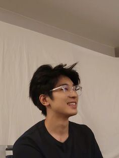 a young man with glasses sitting in front of a white wall holding a nintendo wii controller