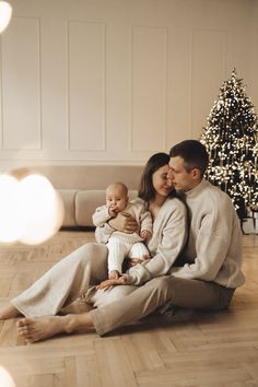 a man and woman are sitting on the floor with their baby in front of a christmas tree