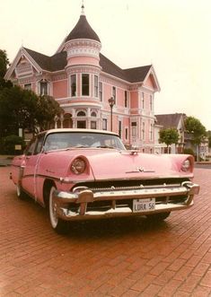 an old pink car is parked in front of a large pink house on a brick street