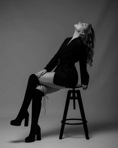 a woman sitting on top of a stool wearing high heel boots and black dress with long sleeves