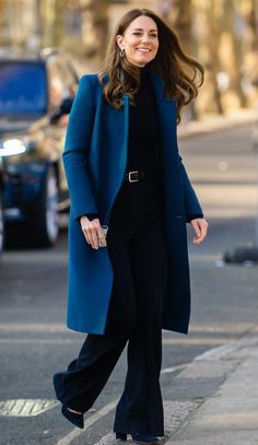 a woman walking down the street wearing a blue coat and black pants with her hands in her pockets