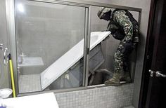 a man in camouflage standing next to a bathtub and sink with the door open