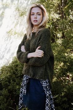 a woman standing with her arms crossed wearing a green sweater and plaid skirt in front of trees