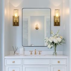 a white bathroom with gold fixtures and flowers on the sink countertop, along with a large mirror above it