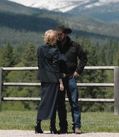 a man and woman standing next to each other in front of a fence with mountains in the background