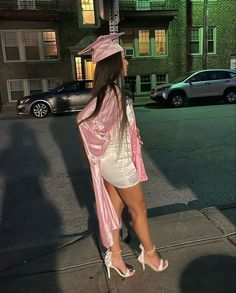 a woman in a pink graduation gown is standing on the sidewalk with her legs crossed