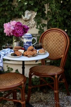 two chairs and a table with food on it in front of some bushes, flowers