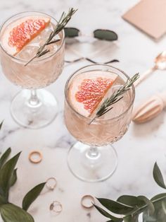 two glasses filled with gin and garnish on top of a marble countertop