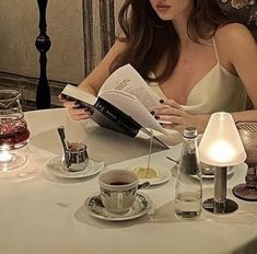 a woman sitting at a table reading a book and drinking tea or coffee with her eyes closed