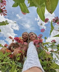 two women are taking a selfie with flowers in the foreground and clouds in the background