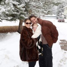 a man, woman and baby are standing in the snow near a tree with their arms around each other