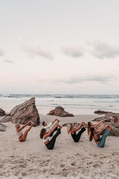 four women doing yoga on the beach in front of some large rocks and water at sunset