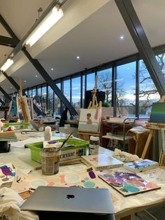 an artist's studio with lots of paintings and art supplies on the table in front of large windows