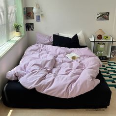 an unmade bed with pink sheets and black pillows in a white room next to a window