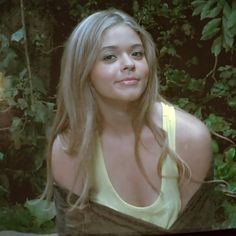 a woman with long blonde hair standing in front of some trees and bushes looking at the camera