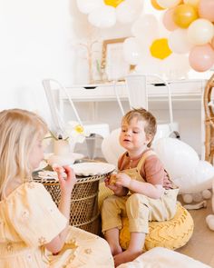 two young children sitting on the floor eating cake and balloons in the air behind them