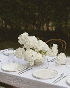 white flowers are in a vase on a table set with plates and silverware for dinner