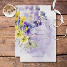 the wedding card is decorated with watercolor pansies and white ribbon on a wooden table