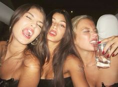 three beautiful young women sitting next to each other holding wine glasses in their mouths and laughing