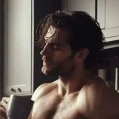 a shirtless man sitting in a kitchen next to a window holding a coffee cup