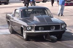 an old muscle car is driving down the street with people walking around in the background