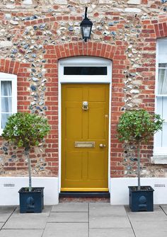 two potted trees in front of a yellow door on a brick building with white windows