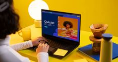 a woman sitting in front of a laptop computer on top of a yellow table next to a lamp