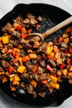 a skillet filled with meat and vegetables on top of a marble countertop next to a wooden spoon