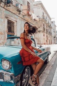 a woman in a red dress leaning on the hood of an old car and smiling