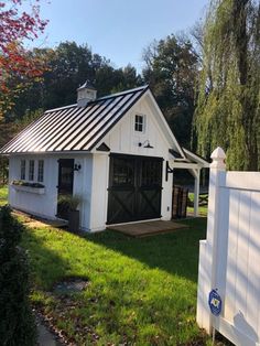 a small white barn with a black roof and door on the grass next to a fence