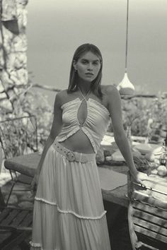 a woman in a white dress standing on a wooden bench next to the ocean and looking at the camera