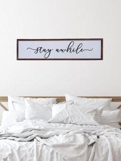 a bed with white sheets and pillows in front of a framed sign that says stay awhile