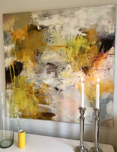 two candles are sitting on a table in front of a large painting with yellow and grey colors