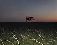 a house in the middle of a field with tall grass at night, lit up by lights