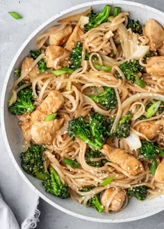 a white bowl filled with noodles, broccoli and chicken on top of a table