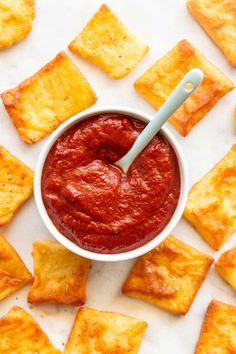 a white bowl filled with red sauce surrounded by crackers on a white counter top